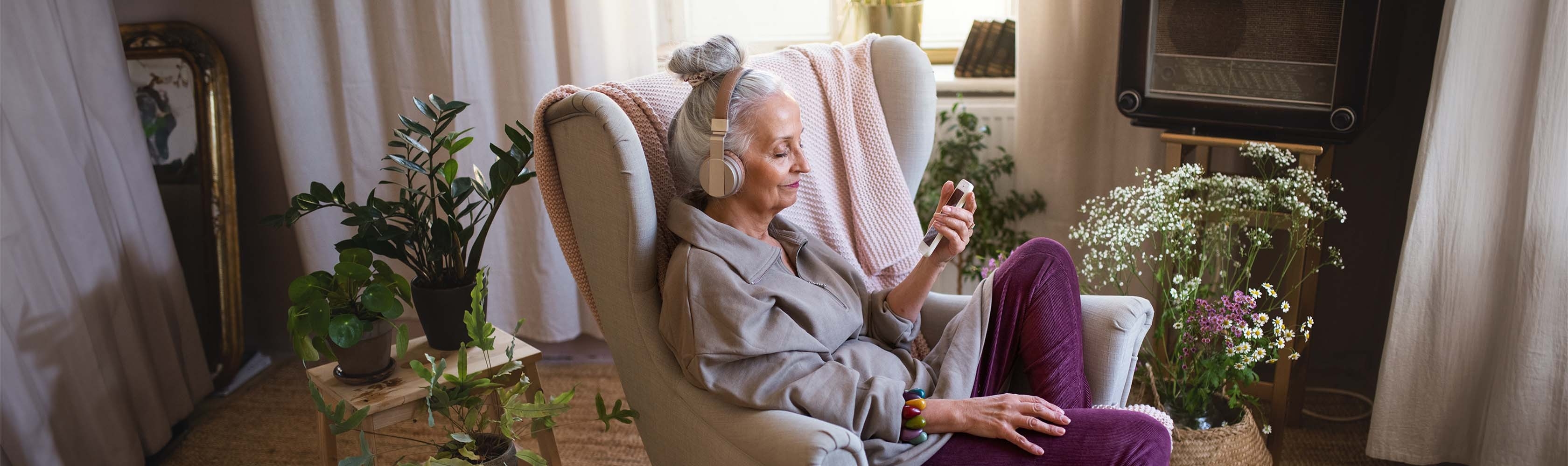 Ältere Frau sitzt auf einem Sessel in ihrem Wohnzimmer und hört einen Podcast zu Gesundheitsthemen 